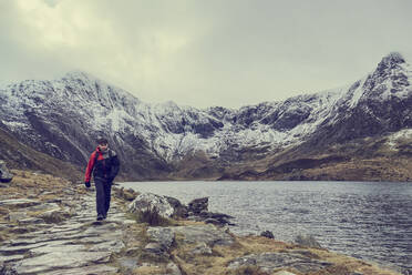 Männlicher Wanderer wandert entlang einer Seenlandschaft mit schneebedeckten Bergen, Llanberis, Gwynedd, Wales - CUF52859