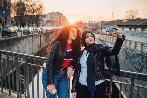 Junge Frau im Hidschab und beste Freundin nehmen Selfie auf Kanalbrücke in der Stadt bei Sonnenuntergang - CUF52876