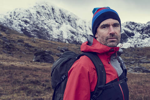 Männlicher Wanderer mit Strickmütze vor schneebedeckten Bergen, Porträt, Llanberis, Gwynedd, Wales, lizenzfreies Stockfoto