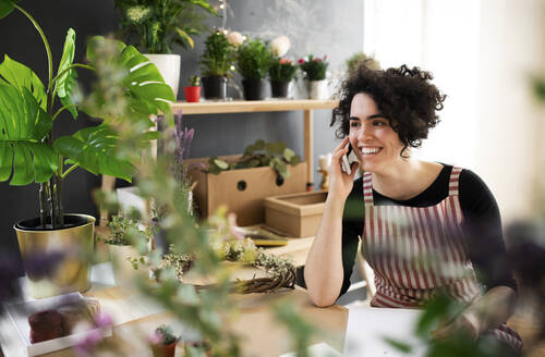 Glückliche junge Frau am Telefon in einem kleinen Laden mit Pflanzen - HAPF03013