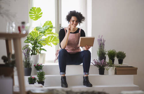 Smiling young woman with cardboard box in a small shop with plants - HAPF03022