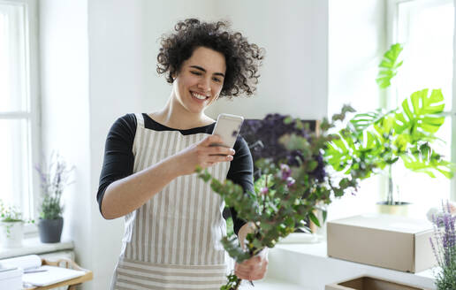 Lächelnde junge Frau, die mit ihrem Smartphone ein Foto von Blumen in einem kleinen Laden macht - HAPF03047