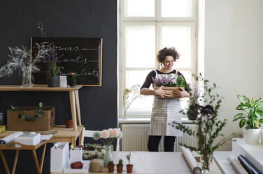 Smiling young woman holding flowers in a cardboard box in a small shop - HAPF03050