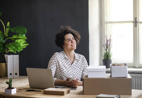 Lächelnde junge Frau sitzt am Schreibtisch mit Kaffeetasse, Laptop und Paketen - HAPF03081
