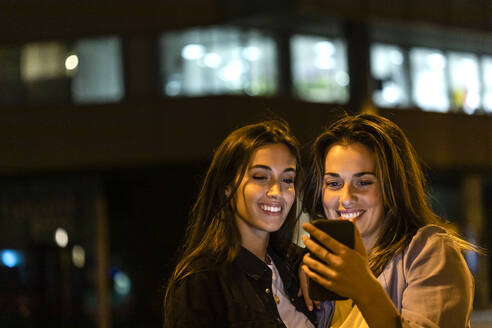 Zwei Freunde benutzen das Smartphone bei Nacht, mit den Lichtern der Stadt im Hintergrund - JRFF03819