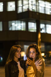 Zwei Freunde benutzen das Smartphone bei Nacht, mit den Lichtern der Stadt im Hintergrund - JRFF03820