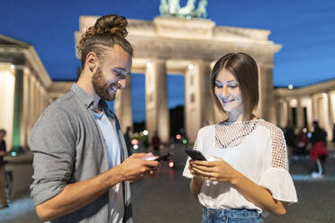 Glückliches Paar mit Smartphones am Brandenburger Tor zur blauen Stunde, Berlin, Deutschland - WPEF02136