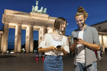 Glückliches Paar mit Smartphones am Brandenburger Tor bei Nacht, Berlin, Deutschland - WPEF02139