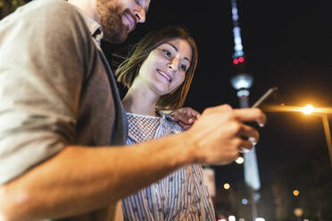 glückliches Paar bei der Nutzung von Smartphones in der nächtlichen Stadt, im Hintergrund der Fernsehturm, Berlin, Deutschland - WPEF02147