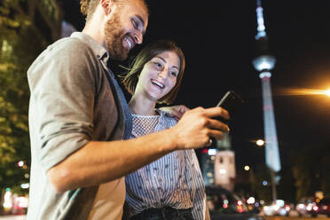 glückliches Paar bei der Nutzung von Smartphones in der nächtlichen Stadt, im Hintergrund der Fernsehturm, Berlin, Deutschland - WPEF02148