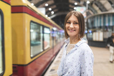 Junge lächelnde Frau auf dem Bahnhof mit verschwommenem Zug im Hintergrund - WPEF02153