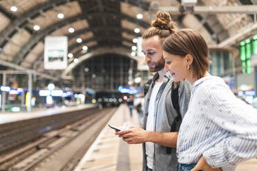 Junges Paar, das am Bahnhof auf den Zug wartet und ein Smartphone benutzt, Berlin, Deutschland - WPEF02154