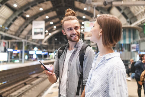 Junges Paar, das am Bahnhof auf den Zug wartet und ein Smartphone benutzt, Berlin, Deutschland, lizenzfreies Stockfoto