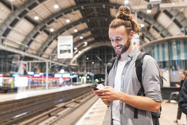 Junger lächelnder Mann mit Smartphone auf dem Bahnhof - WPEF02157