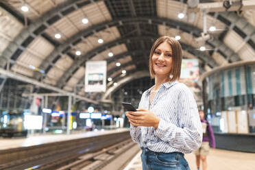 Junge lächelnde Frau mit Smartphone auf dem Bahnhof - WPEF02159