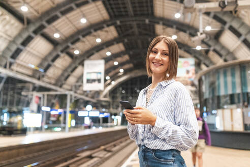 Junge lächelnde Frau mit Smartphone auf dem Bahnhof - WPEF02159