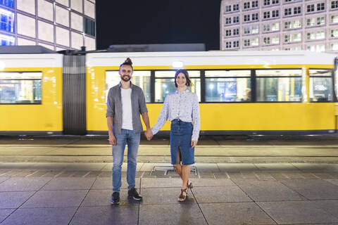 Glückliches Paar, das sich an den Händen hält und in die Kamera schaut, mit Straßenbahn im Hintergrund, Berlin, Deutschland, lizenzfreies Stockfoto