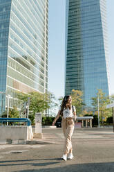 Young woman with take away drink crossing road, high-rise buildings in the background - KIJF02667