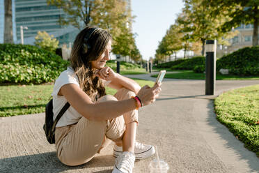 Young woman sitting in park using smartphone and listening to music with headphones - KIJF02672