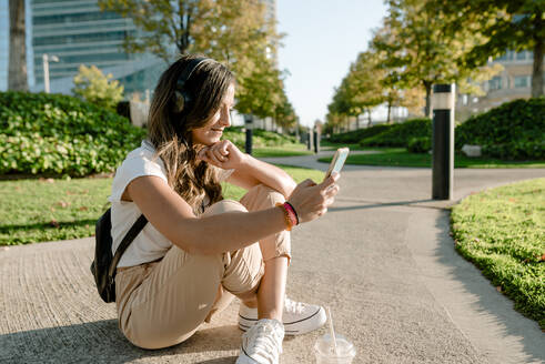 Junge Frau, die im Park sitzt, ihr Smartphone benutzt und mit Kopfhörern Musik hört - KIJF02672