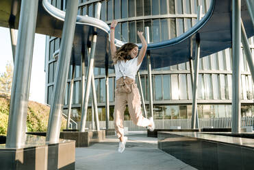 Young woman with headphones jumping in business area - KIJF02674