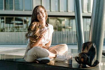 Young smiling woman sitting in business area with hamburger and milkshake, looking at camera - KIJF02676