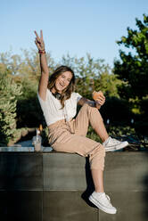 Young smiling woman with hamburger and milkshake raising hand in victory sign - KIJF02681