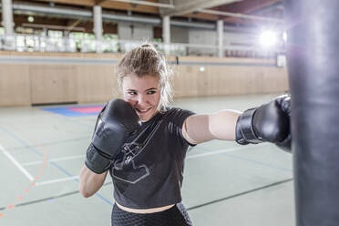 Smiling female boxer practising at punchbag in sports hall - STBF00460