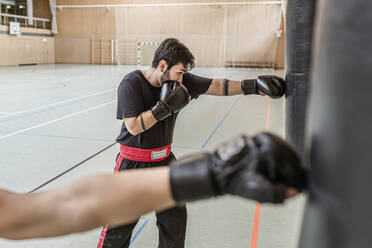 Coach and boxer practising at punchbags in sports hall - STBF00464