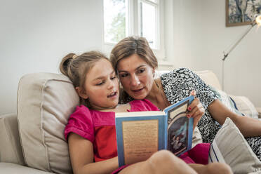 Mother reading book with daughter on couch in living room - EGBF00368