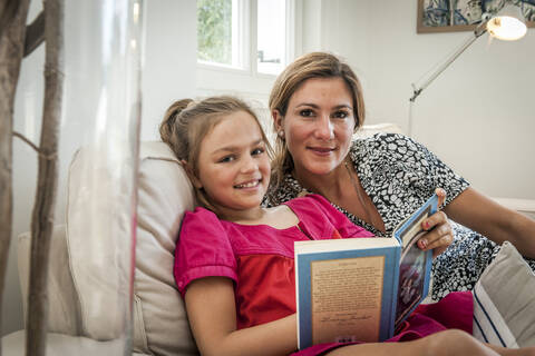 Porträt einer lächelnden Mutter und Tochter mit Buch auf der Couch im Wohnzimmer, lizenzfreies Stockfoto