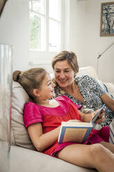 Happy mother and daughter with book on couch in living room - EGBF00370