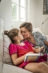 Mother kissing daughter with book on couch in living room - EGBF00371
