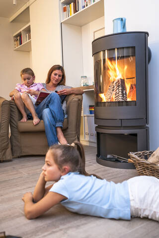 Mutter mit Töchtern entspannt und liest im Wohnzimmer am Kamin, lizenzfreies Stockfoto