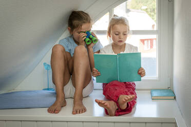 Mädchen mit Spielzeug-Fernglas schaut auf Schwester mit Buch am Fenster - EGBF00458