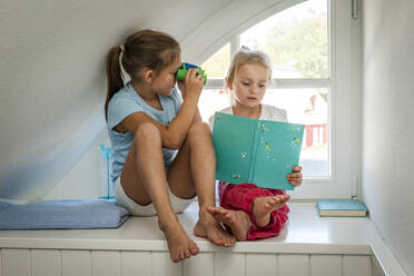 Mädchen mit Spielzeug-Fernglas schaut auf Schwester mit Buch am Fenster - EGBF00459