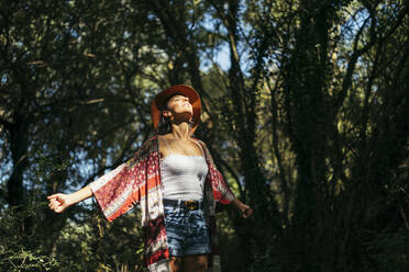 Young woman wearing a brown hat, colorful shirt and white top with closed eyes and arms up feeling the sun in a forest - MTBF00049