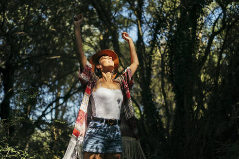 Junge Frau mit braunem Hut, buntem Hemd und weißem Oberteil, mit geschlossenen Augen und erhobenen Armen, die die Sonne in einem Wald spüren - MTBF00050