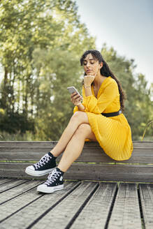 Junge Frau in gelbem Kleid und schwarzen Turnschuhen, die auf einer Holzpromenade ihr Smartphone benutzt - MTBF00054