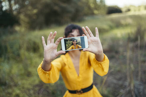 Glückliche junge Frau im gelben Kleid, die ein weißes Smartphone vor ihr Gesicht hält und ein Selfie macht - MTBF00055