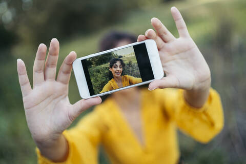 Glückliche junge Frau im gelben Kleid, die ein weißes Smartphone vor ihr Gesicht hält und ein Selfie macht, lizenzfreies Stockfoto