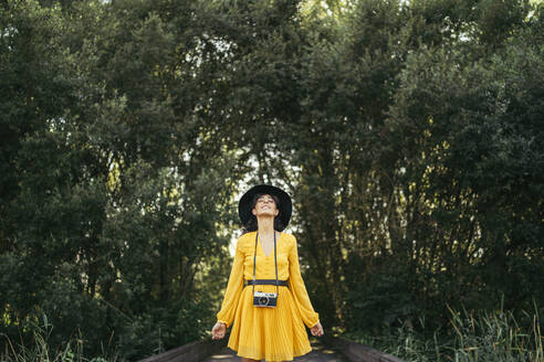 Junge Frau mit schwarzem Hut und gelbem Kleid mit einer analogen Kamera auf einer Holzpromenade - MTBF00057