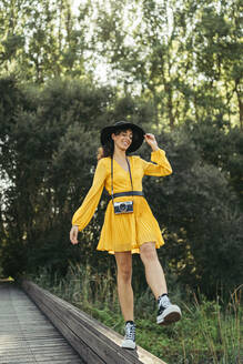 Junge Frau mit schwarzem Hut und gelbem Kleid mit einer analogen Kamera auf einer Holzpromenade - MTBF00061