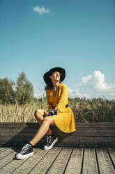 Young woman wearing a black hat and yellow dress with an analog camera sitting on wooden boardwalk - MTBF00062