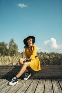 Young woman wearing a black hat and yellow dress with an analog camera sitting on wooden boardwalk - MTBF00062
