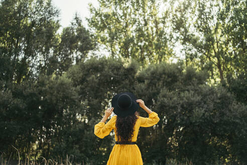 Rückansicht einer jungen Frau mit schwarzem Hut und gelbem Kleid, die die Natur genießt - MTBF00063