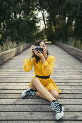 Junge Fotografin in gelbem Kleid und schwarzen Turnschuhen hält eine analoge Kamera auf einer Holzpromenade - MTBF00065