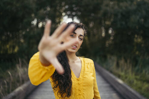 Junge Frau mit schwarzem Hut und gelbem Kleid mit einer analogen Kamera schüttelt die Hand zu einer Kamera auf der Holzpromenade - MTBF00066