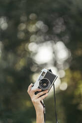 Hand holding grey and black analog camera with forest bokeh effect in background - MTBF00067