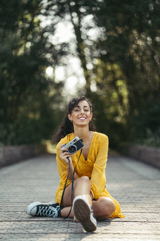 Junge Fotografin in gelbem Kleid und schwarzen Turnschuhen hält eine analoge Kamera auf einer Holzpromenade, lizenzfreies Stockfoto